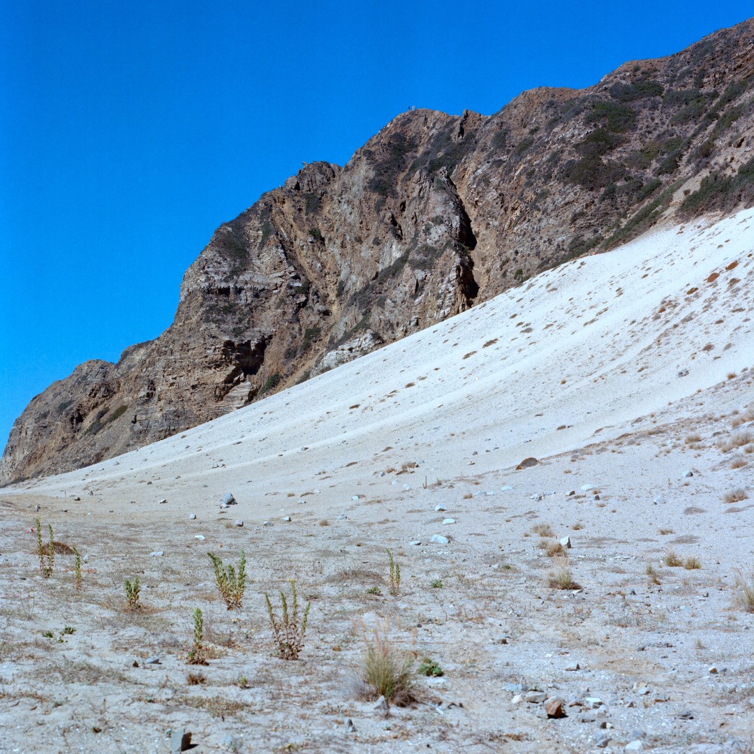 Sandy slope next to a beach with white sand occupying bottom half of photo and rocky outcrop running in the background and a blue sky.