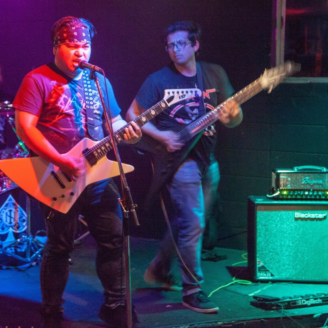 Photo of metal band playing on stage at a bar with lead singer on guitar and bassist visible and drummer obscured. Color scheme is red, violet and blue lights