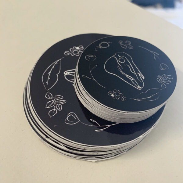 Photo of a stack of smaller stickers on top of a stack of larger stickers. Both have the same design showing a horse skull in the middle and various items such as nuts, flowers and seashells all in white on a black background