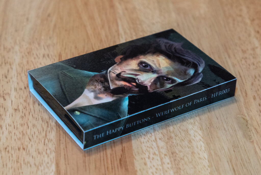 Photo of cassette single on wooden counter. Name Werewolf of Paris, as well as album cover featuring person with crazed look on his face and dark graveyard background can be seen facing up