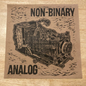 Photo of black print on brown paper that says "NON-BINARY ANALOG" with a giant scorpion on top of a view camera